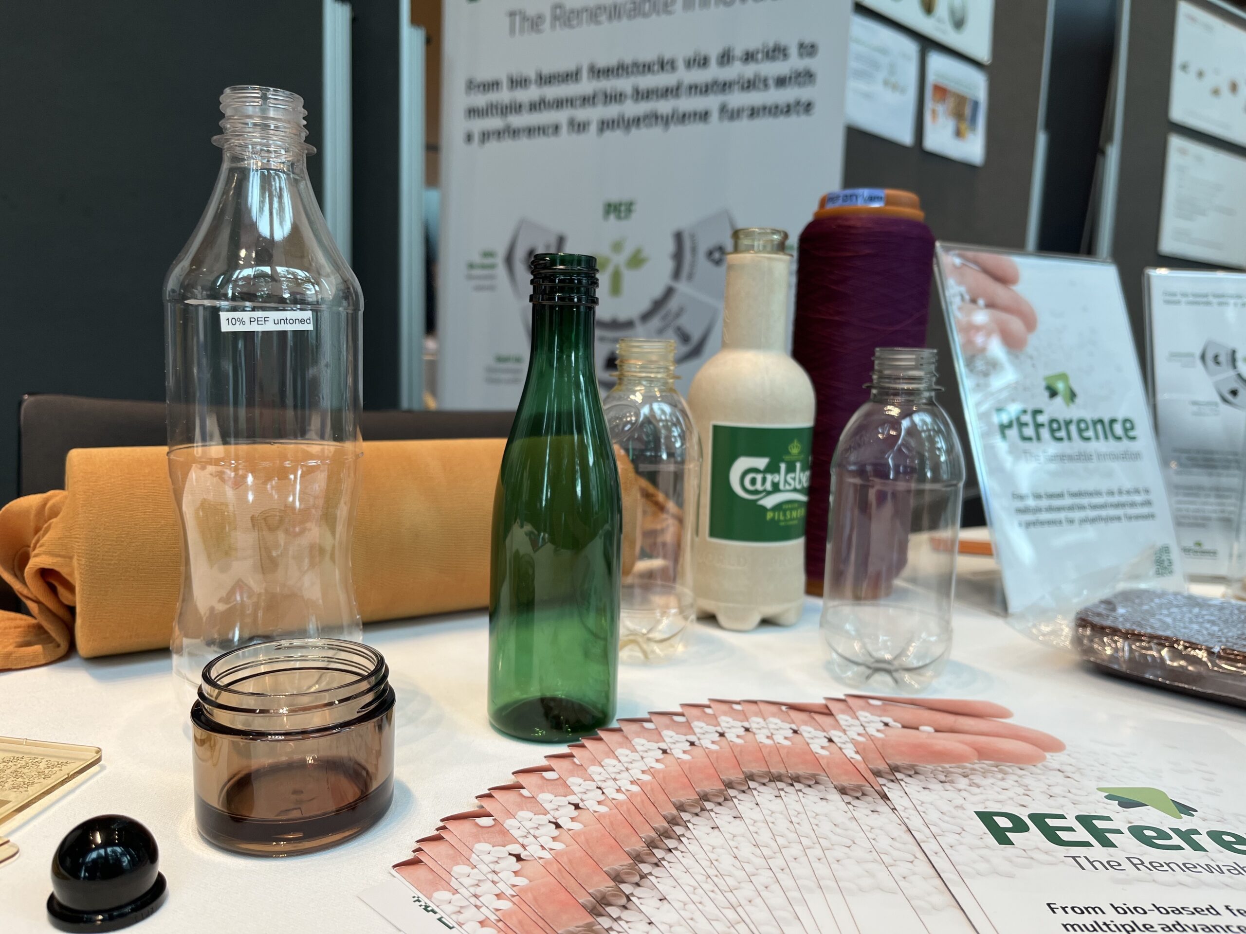 PEF bottles (clear and green), Carlsberg paper bottle, PEFerence flyers, see-through grey-black cosmetics crucible on a white table, PEFerence roll-up in the background