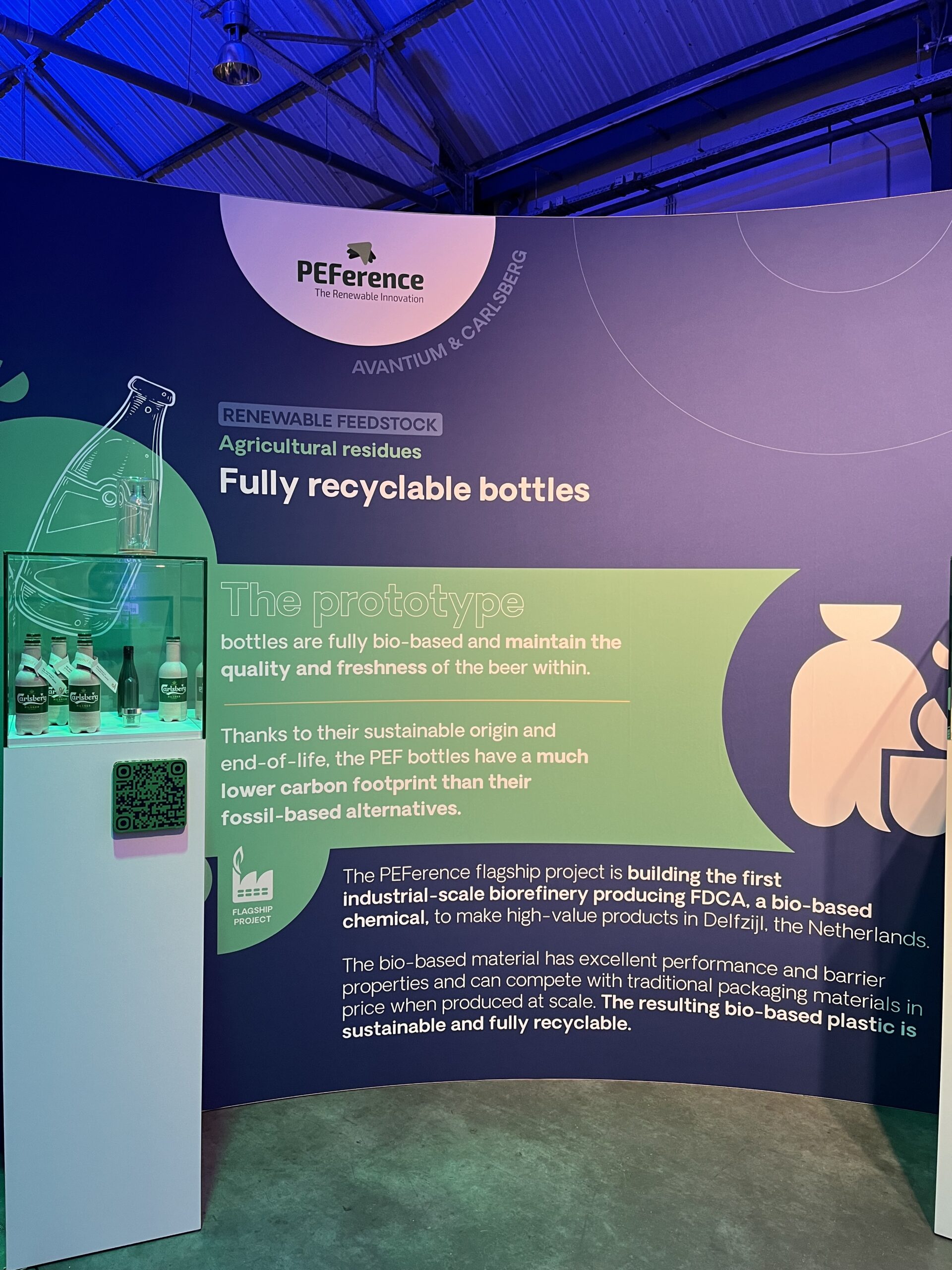 Carlsberg paper bottle and PEF bottles displayed in glass showcase, PEFerence logo and text about demonstrators at a green and violet curved wall in the background, violet and green lights 