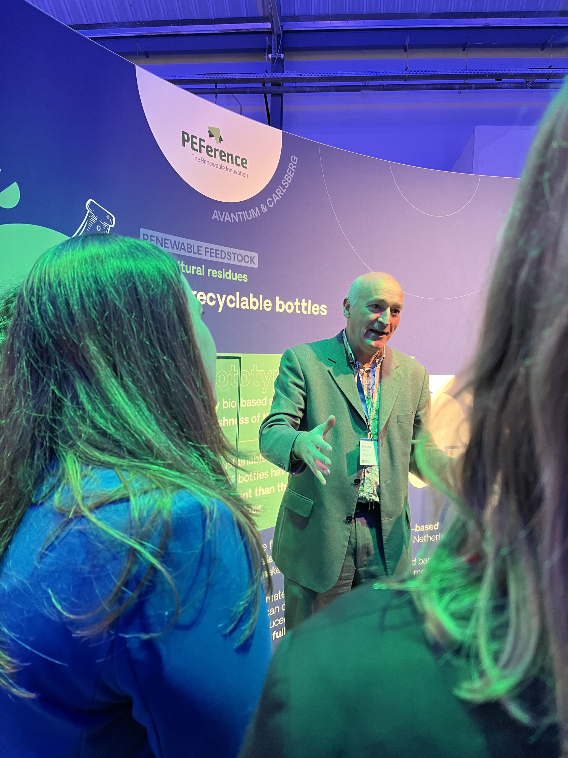 Ed de Jong opens the PEFerence demonstrator display to an interested audience. Green and violet lights. Two long-haired persons visible in the front.