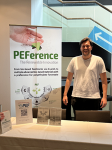 The PEFerence booth at the Renewable Materials Conference 2024. In front is a table with information material and on the left side behind the table is a roll-up banner of the project. On the right side behind the table is Kristijan Mrsic from nova-institute, one of the representatives of PEFerence.